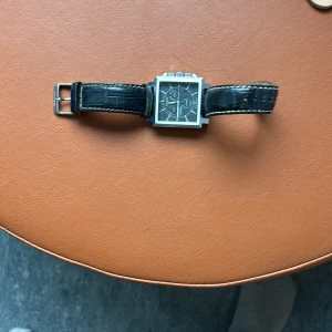 Svart fyrkantig klocka med läderband - Stilren klocka med fyrkantigt silverfärgat boett och svart urtavla. Klockan har svart läderarmband med vita sömmar och flera urtavlor för extra funktioner. Perfekt accessoar för dig som gillar en modern och klassisk look.