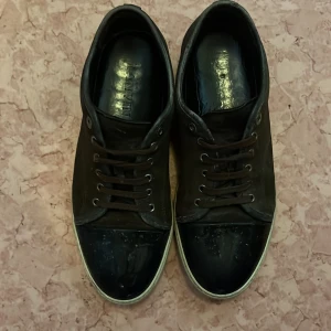 Svarta sneakers från Lanvin i skinn - Snygga svarta sneakers från Lanvin med klassisk vit sula och rund tå. Ovandelen är i skinn och tån är i blankt material för en cool kontrast. Skorna har bruna snören och diskreta metalldetaljer vid snörningen. Perfekta för dig som gillar stilrena och lyxiga sneakers.
