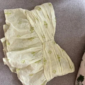 Gul broderad bandeau tubtopp - Supersöt ljusgul bandeau tubtopp i linne med broderade gröna detaljer. Toppen har volangig form och är perfekt för varma dagar. Sitter snyggt axelbandslöst och har en luftig känsla.
