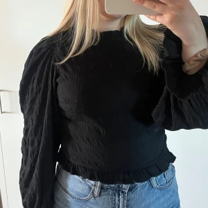 Svart blus med volang från Bubbleroom - Supersnygg svart blus från Bubbleroom med volangdetaljer vid halsringning och ärmslut. Blusen har långärmade puffärmar.