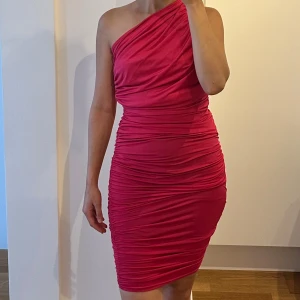 Rosa one shoulder kortklänning - Säljer en tight rosa kortklänning med one shoulder-design och snygga rynkade detaljer. Klänningen har en figurnära passform och är perfekt för dig som vill sticka ut. Materialet känns mjukt och följsamt mot kroppen.