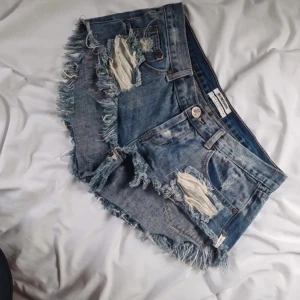 Blå slitna jeansshorts från One Teaspoon low waist  - Riktigt snygga blå jeansshorts från One Teaspoon med slitna detaljer, fransig kant och klassisk femficksmodell. Shortsen har låg midja och är perfekta för sommaren. Rå och avslappnad vibe med coola trasiga partier och slitningar.