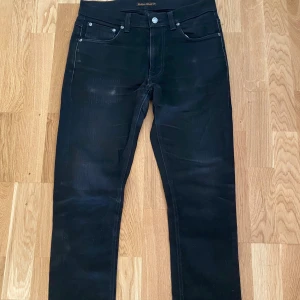 Svarta jeans från Nudie Jeans str:31 - Säljer ett par svarta jeans från Nudie Jeans med klassisk femficksdesign och diskreta svarta sömmar på bakfickorna. Jeansen har raka ben och normal passform, tillverkade i mjukt denimtyg. Perfekta för en clean och stilren look.