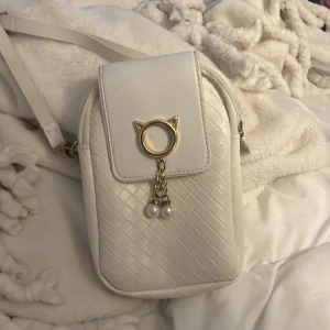 Vit axelväska med pärldetaljer - Söt vit axelväska med flätad struktur och justerbar rem. Locket har en unik guldfärgad kattöronsring med hängande pärlor som ger väskan en lekfull touch. Perfekt storlek för mobilen och småsaker, och stängs med magnetknapp.
