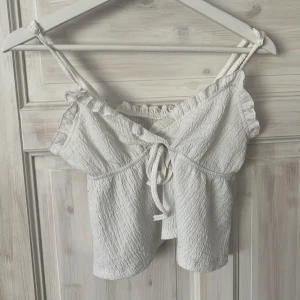 Vit croppad linnetopp från SHEIN - Supersöt vit croppad linnetopp från SHEIN med smala axelband, volangkant och knytning framtill. Toppen har en lätt skrynklig struktur och är perfekt för varma dagar. Enkel att matcha med jeans eller kjol för en chill look.