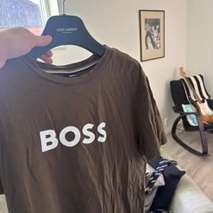 Brun BOSS t-shirt M - Snygg brun t-shirt från BOSS med vit logga tryckt på bröstet. Klassisk rund halsringning och korta ärmar. Perfekt för dig som gillar stilrena och enkla plagg med tydlig branding. Tillverkad i mjuk bomull för skön känsla hela dagen.