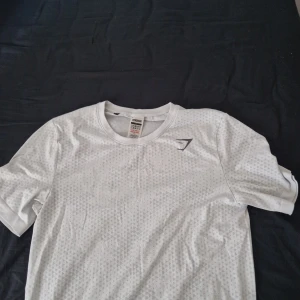 Vit Gymshark t-shirt med mönster - Säljer en vit t-shirt från Gymshark med diskret geometriskt mönster över hela tyget och logga på bröstet. T-shirten är kortärmad och har en sportig vibe, perfekt för träning eller chill. Materialet är mjukt och andas bra.