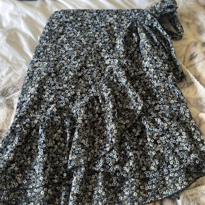 Blommig omlottkjol från ONLY - Supersöt svart omlottkjol från ONLY med små blå och vita blommor. Kjolen har volangkant och knytband i midjan för en snygg passform. Perfekt för dig som gillar romantiska och trendiga vibes. Dock så är öglan där man drar igenom snöret trasigt men går att klippa bort eller sy dit