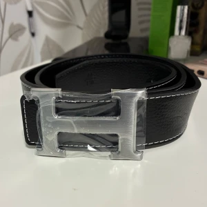 Svart skärp med H-spänne - Stilrent svart skärp i skinn med en stor, silverfärgad H-formad metallspänne. Bältet har vita kontrastsömmar längs kanterna som ger en snygg detalj. Perfekt accessoar för att lyfta vilken outfit som helst. 