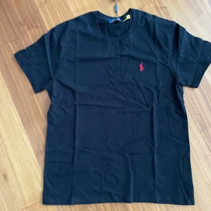 Svart Polo Ralph Lauren T-shirt  - Polo Ralph Lauren T-shirt Storlek L Svart med röd logga Helt ny – aldrig använd  Klassisk och helt ny t-shirt från Polo Ralph Lauren – aldrig använd. Perfekt för en avslappnad men stilfull look tack vare den tidlösa designen. Ikonisk logga broderad i rött på bröstet. Kommer med originaletiketter. Ett måste i garderoben – passa på! Pris kan diskuteras. Skriv om du vill ha fler bilder!