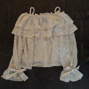 Vit spetsblus med pärlband från ROJITA - Superfin vit blus i spets från ROJITA, perfekt för jirai kei/ryousangata, med volanger och pärlprydda axelband. Blusen har dekorativa pärlknappar framtill, långa ärmar med breda manschetter och stora rosetter. Ena ”stenen” på lägsta knappen är borta men kan enkelt ersättas. Finns liknande att hitta på Aliexpress.