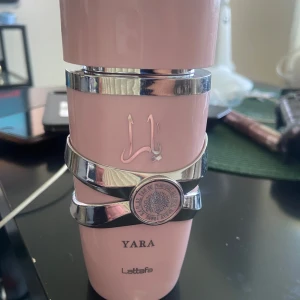 Yara parfym från Lattafa - Säljer en snygg parfymflaska Yara från Lattafa i ljusrosa med silvriga detaljer och dekorativa band runt flaskan. Flaskan har en modern design och arabiska bokstäver på framsidan. Perfekt för dig som gillar unika och trendiga dofter. Tagit några sprut. Nästan full.