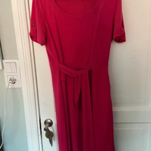 Mörkrosa Esprit klänning med knytband - Snygg mörkrosa klänning från Esprit med rund halsringning och knytband i midjan. Modellen har korta ärmar med knappdetalj och är tillverkad i ett mjukt, bekvämt material. Perfekt för dig som gillar enkel men stilren design. Nytt skick!