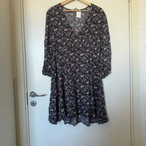 Mörkblå blommig kortklänning med v-ringning - Supersöt svart kortklänning med v-ringning och trekvartsärm. Klänningen har ett blommigt mönster i rosa, lila och beige samt små solfjädrar. Den är luftig och har lätt volang nertill, perfekt för vår och sommar. Från märket Odd Molly i storlek 2 som enligt deras storleksguide motsvarar en storlek M. Fint skick, men tyvärr finns tråddraningar. 