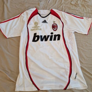 AC Milan vit bortatröja Adidas - AC Milan bortatröja i vitt med svarta och röda detaljer, Adidas-logga och Bwin sponsortryck. Champions League-finalen 2007 broderat i guld på bröstet. Kortärmad modell i lätt och ventilerande material, perfekt för fotbollsälskare.