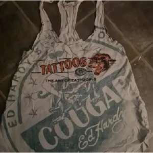 Y2k Ed Hardy linne  - Coolt y2k linne från Ed Hardy med stort tattoo-inspirerat tryck i grönt, orange och svart. Framsidan har texten 'TATTOOS' och 'COUGAR', samt klassiska Ed Hardy-motiv. Baksidan har flames och yin-yang-symbol. Tunt material och brottarrygg.  Inköpt i USA någon gång under 2000-talet. 