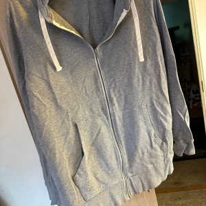 Grå hoodie från Jack & Jones strl: XXL - Säljer en grå hoodie från Jack & Jones med dragkedja framtill, huva med vita snören och två stora fickor. Tröjan är i mjuk bomullsmix och har ribbade muddar vid ärmslut och nederkant. Perfekt för chill dagar eller när du vill ha en enkel och clean look.