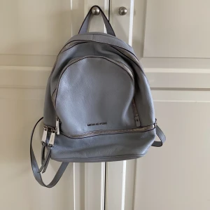 Grå ryggsäck från Michael Kors - Snygg grå ryggsäck från Michael Kors i mjukt skinn. Väskan har flera dragkedjefack, justerbara axelremmar och silverfärgade detaljer. Mycket fint skick, inga skavanker eller andra anmärkningar. Från Michael Kors. 