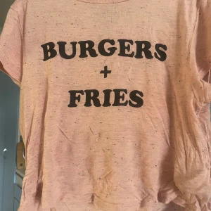 Rosa t-shirt med text Burgers + Fries - Rosa t-shirt med svart tryck där det står 'BURGERS + FRIES' på framsidan. T-shirten har korta ärmar och är tillverkad i ett mjukt material med små svarta prickar i tyget. Perfekt för dig som gillar chill och streetstyle.