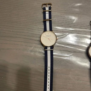 Randig klocka från Winchester - Stilren klocka från Winchester med vit urtavla och tunna guldiga detaljer. Armbandet är randigt i marinblått och vitt, gjort i slitstarkt tygmaterial. Perfekt accessoar för en clean och klassisk look. Klockan är helt ny i kartong och är aldrig använd 