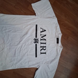 Vit AMIRI t-shirt med logga - Snygg vit t-shirt från AMIRI med stor svart logga och grafisk detalj framtill. Klassisk rund halsringning och korta ärmar. Tillverkad i mjuk bomull för en skön känsla och clean look. Perfekt för dig som gillar streetwear och vill sticka ut med ett exklusivt märke.