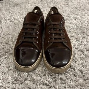 Bruna sneakers från Lanvin i mocka - Snygga bruna sneakers från Lanvin med ovandel i mocka och lackad tå. Perfekt för dig som gillar stilrena och lyxiga detaljer i vardagen. Mycket bra skick, mindre skada på sulan