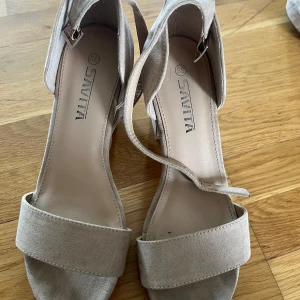 Beige sandaler med klack från Savita storlek 38 - Snygga beige sandaler från Savita med bred rem över tårna och justerbart spänne vid ankeln. Skorna har en stadig blockklack och är tillverkade i mockaliknande material. Perfekta för dig som gillar stilrena och trendiga skor med öppen tå. storlek 38
