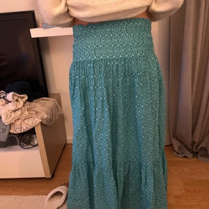 Turkos långkjol med mönster - Säljer en turkos långkjol med elastisk smock i midjan och coolt geometriskt mönster. Kjolen är luftig och har en lätt volang nertill, perfekt för dig som gillar en avslappnad och trendig stil. Den är i M men passar perfekt på mig som har storlek xxs-xs i jeans då den har en väldigt stretchig midja.