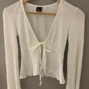 Vit omlottopp från Gina Tricot XS - Supersöt vit omlottopp från Gina Tricot i storlek XS. Toppen har långa ärmar, v-ringning och knyt framtill. Materialet är lätt och vågigt med struktur, vilket ger en luftig och trendig känsla. Perfekt att styla med jeans eller kjol för en fräsch look. Använd en gång.