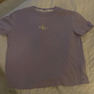 Lila t-shirt från Calvin Klein Jeans - Säljer en lila t-shirt från Calvin Klein Jeans med klassisk logga i gult och vitt på bröstet. T-shirten är i mjuk bomull och har en enkel, rak passform med korta ärmar. Perfekt för dig som gillar stilrena och bekväma plagg.