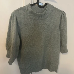 Grön stickad tröja med puffärm - Mysig grön stickad tröja med bred ribbad krage och puffiga trekvartsärmar. Tröjan har en avslappnad passform och ribbade muddar vid ärmslut och nederkant. Perfekt för lager-på-lager under kyliga dagar. 