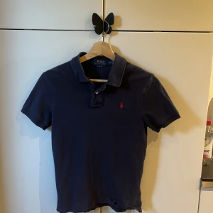 Marinblå pikétröja Polo Ralph Lauren - Snygg marinblå pikétröja från Polo Ralph Lauren i slim fit-modell. Klassisk krage, korta ärmar och röd broderad logga på bröstet. Tillverkad i mjuk bomull, perfekt för en clean och stilren look.
