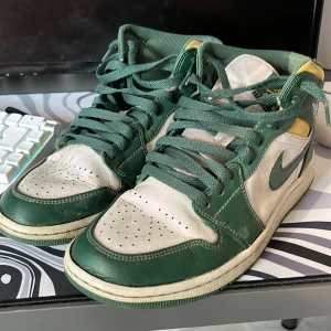 Nike Air Jordan 1 gröna sneakers - Nike Air Jordan 1 sneakers i grönt och vitt med gula detaljer. Klassisk high-top modell med snörning, perforerad tå och ikonisk Jumpman-logga på plösen. Ovandel i skinn och textil, perfekt för dig som gillar retrostil och streetwear. Såklart äkta