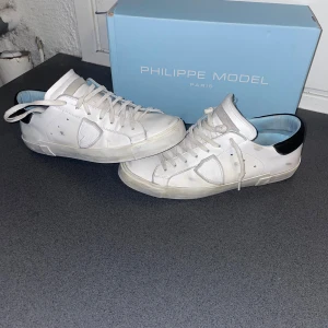 Vita Philippe Model sneakers i skinn - Snygga vita sneakers från Philippe Model Paris med svarta detaljer på hälen. Skorna har klassisk låg profil, rund tå och snörning. Tillverkade i skinn med vit sula och diskret logga på sidan. Perfekta för en clean och stilren look.