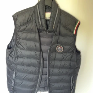Svart dunväst från Dolomite XL - Snygg svart dunväst från Dolomite med quiltad design och dragkedja framtill. Västen har två fickor med dragkedja och en broderad logga på bröstet. Detalj med italienska flaggans färger på axeln ger en sportig touch.