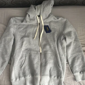 Grå hoodie från Polo Ralph Lauren - (Varan är helt ny )Snygg ljusgrå hoodie från Polo Ralph Lauren med dragkedja framtill, vita snören i huvan och två stora fickor. Tillverkad i mjuk bomullsmix som känns riktigt skön. Perfekt för dig som gillar en clean och klassisk stil.
