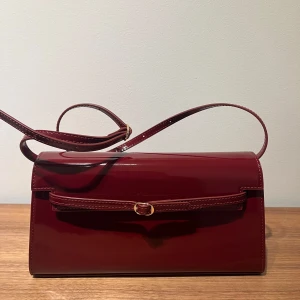 Röd väska från Verafied - Snygg vinröd axelväska i patent leather från Verafied. Väskan har en elegant design med ett spänne framtill och en justerbar axelrem, kan användas som clutch, handväska eller axelremsväska. Limited edition. Ursprungspris 2907 kr. Nyskick aldrig använd. 