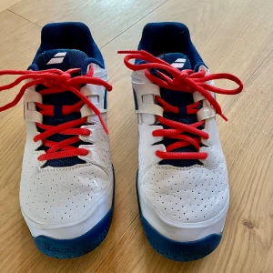 Tennis/-paddelskor junior - Tennis-/Padelskor från Babolat för junioren. Storlek 38,5. Vit med blå detaljer.  Sko som erbjuder stabilitet, hållbarhet och komfort, med en rem över mellanfoten, ett skyddande stycke för framfoten och Michelinsula designad för att hålla på alla typer av underlag.  Använda i tennishallen under en säsong.