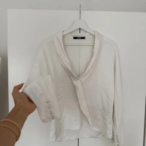 Vit långärmad skjorta från Bik Bok - Säljer en stilren vit skjorta från Bik Bok i storlek XS. Skjortan har lång ärm med knappar vid ärmslut, en snygg v-ringad krage och är tillverkad i ett mjukt, lätt material som ger en lyxig känsla. Perfekt för dig som gillar minimalistisk och clean stil.