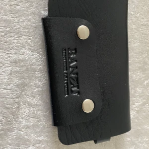 Svart nyckelplånbok  - Snygg svart nyckelplånbok från BANZU med silverfärgade detaljer. Plånboken har flera krokar för nycklar och tryckknappar för smidig stängning. Perfekt att ha i fickan eller väskan för att hålla koll på nycklarna. Finns brun och svart färger kvar 