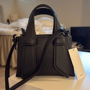Svart handväska från Mango - Snygg svart handväska från Mango med stilren design och coola detaljer. Väskan har både handtag och axelrem, samt guldfärgad dragkedja upptill. Helt ny, aldrig använd.