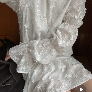 Vit blommig kortklänning med öppen rygg - Superfin vit kortklänning med broderade blommor, puffiga långärmar och öppen rygg med knytband. Klänningen har volangdetaljer och är gjord i ett tunt, luftigt material. Klänningen är köpt på Nakd och användts 1 gång på midsommar. 