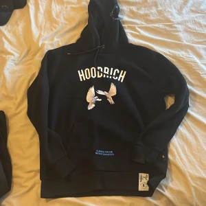 Svart hoodie från Hoodrich med tryck - Svart hoodie från Hoodrich med stor vit logga och duvor tryckta på både fram- och baksidan. Snygga blå detaljer och texten 'From Nothing To Something' på ryggen. Klassisk huva med blå logga.  