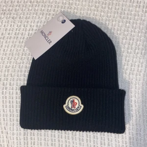 Svart ribbad mössa från Moncler - Säljer en snygg svart ribbad mössa från Moncler. Den är i nytt skick och har en broderad Moncler-logga framtill. Perfekt för att hålla sig varm under höst och vinter. Passar de flesta tack vare one size-storleken. Stilren och enkel att matcha med olika outfits!