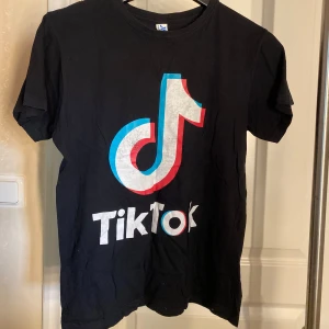 Svart TikTok t-shirt - Svart t-shirt med stort TikTok-logga i vitt, blått och rött framtill. Klassisk rund hals och korta ärmar. Perfekt för dig som älskar sociala medier och vill visa din stil. 