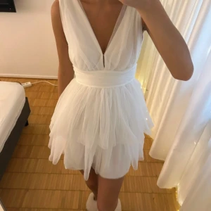 Vit kortklänning med volang och v-ringning - Supersöt vit kortklänning med djup v-ringning och bred midja. Klänningen har flera lager av tunt, skirt tyg som ger en fluffig volang-effekt. Har inga defekter alls!