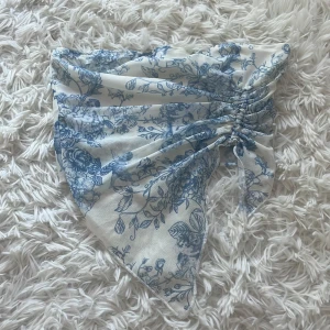 Blommig bandeau tubtopp i blått och vitt - Superfin bandeau tubtopp med blått blommönster på vit botten. Toppen har en spetsig, scarf-liknande nederkant och snygg rynkdetalj framtill med knyt. Perfekt för sommarens alla tillfällen och ger en romantisk vibe.