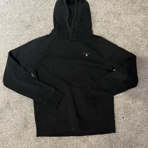Svart hoodie från Gant med huva - Svart hoodie från Gant med klassisk huva och liten broderad logga på bröstet. Tröjan har långa ärmar, känguruficka framtill och är gjord i mjuk bomull. Perfekt för en chill och stilren look. Bara skriva vid frågor och funderingar! STRL 158/164