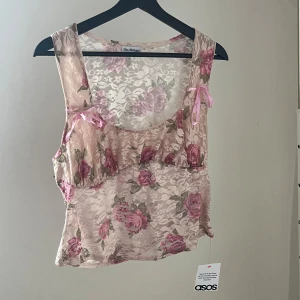 Rosa korsett topp med blommigt tryck  - En jätte söt armlös linne med ljusrosa spets och blommigt mönster. Två rosa rosetter på framsidan. Perfekt nu till sommaren samt för dig som gillar vintagevibbar och mjuka pasteller.