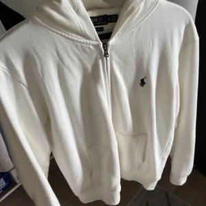Vit hoodie från Polo Ralph Lauren - Snygg vit hoodie från Polo Ralph Lauren med dragkedja framtill och klassisk blå logga på bröstet. Tröjan har huva, långa ärmar och känguruficka. Perfekt för en chill och stilren look.